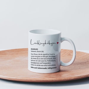 May include: White ceramic mug with a black and white design that reads "Lieblingskollegin [SARAH] Substantiv, feminin (diel Eine Person, die der eigentliche Grund ist, warum man gerne zur Arbeit geht. Eine Kollegin, die immer da ist, wenn man sie braucht. Auch bekannt als Gute-Laune-Fee, Probleml&ouml;serin, Kampfgenossin und Trostspenderin. Ohne sie m&ouml;chte man einfach nicht arbeiten. Siehe auch: Seelenverwandte, Lieblingsmensch"