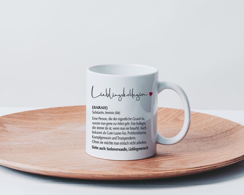 May include: White ceramic mug with a black and white design that reads "Lieblingskollegin [SARAH] Substantiv, feminin (diel Eine Person, die der eigentliche Grund ist, warum man gerne zur Arbeit geht. Eine Kollegin, die immer da ist, wenn man sie braucht. Auch bekannt als Gute-Laune-Fee, Probleml&ouml;serin, Kampfgenossin und Trostspenderin. Ohne sie m&ouml;chte man einfach nicht arbeiten. Siehe auch: Seelenverwandte, Lieblingsmensch"
