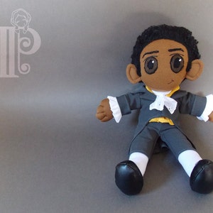 May include: A plush toy of a person with brown skin, black hair, and large brown eyes. The toy is wearing a gray jacket with white ruffles, a white cravat, yellow vest, white socks, and black shoes.