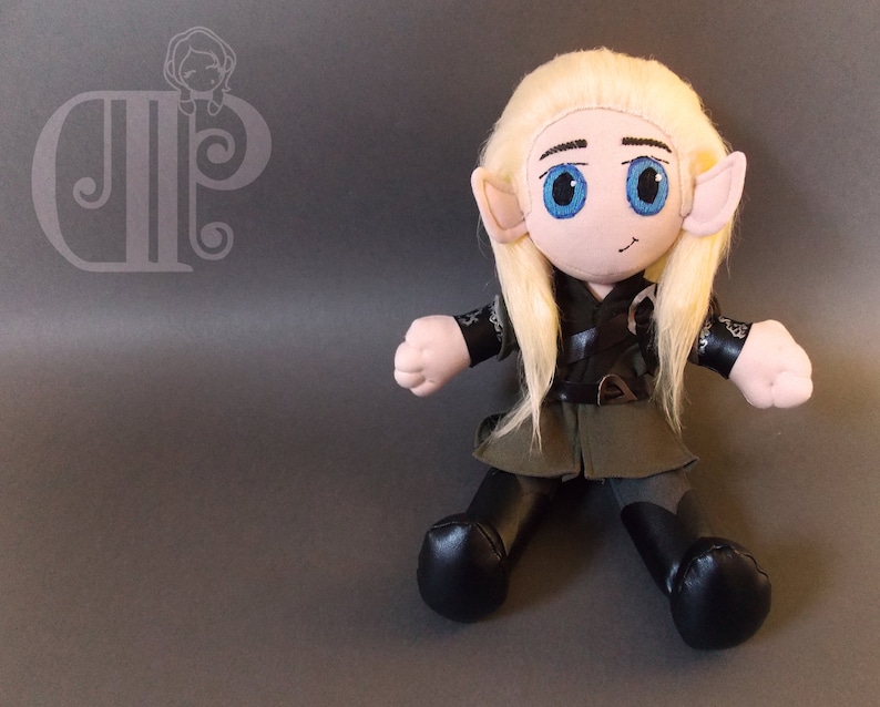 Pode incluir: Um brinquedo de pel&uacute;cia de um elfo com cabelo longo e loiro, vestindo uma roupa verde e preta com um cinto marrom. O elfo tem olhos azuis e um rosto sorridente.