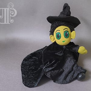 Könnte beinhalten: Eine Plüsch-Hexenpuppe mit schwarzer Samtkleidung und einem spitzen schwarzen Hut. Die Puppe hat grüne Augen, gelbe Haut und schwarze Haare.