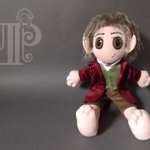 May include: A plush doll of a character from a fantasy novel. The doll has a red jacket, green vest, and brown pants. It has large, expressive eyes, pointed ears, and light brown hair. The doll is sitting with its legs outstretched.