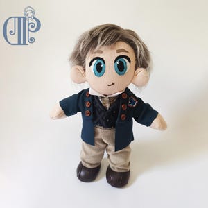 May include: A handmade doll with a stylized design, featuring a navy blue jacket with brown buttons, a black vest, and tan corduroy pants. The doll has light brown hair, large blue eyes, and brown shoes. The doll is standing against a white background.