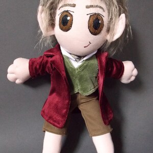 May include: A plush doll depicting a character with light brown hair, large brown eyes, and pointed ears. The doll wears a burgundy jacket, a green vest, brown shorts, and a white collared shirt. The doll's feet are large and rounded.