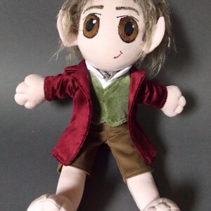 May include: A plush doll depicting a character with large brown eyes, pointed ears, and light brown hair. The doll wears a burgundy jacket, a green vest, a white shirt, and brown shorts. The doll's feet are large and rounded.