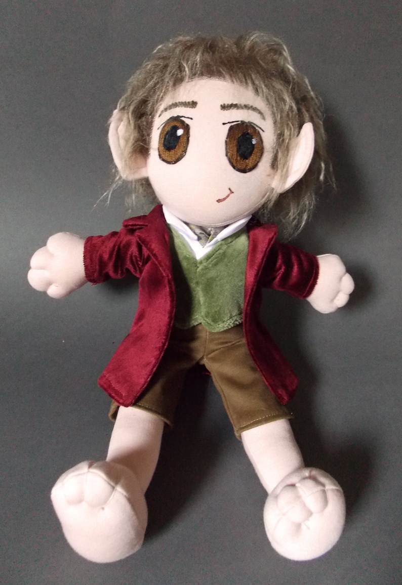 May include: A plush doll depicting a character with large brown eyes, pointed ears, and light brown hair. The doll wears a burgundy jacket, a green vest, a white shirt, and brown shorts. The doll's feet are large and rounded.