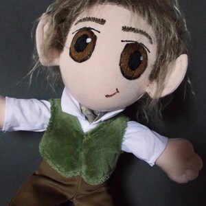 May include: A handmade doll with a fabric face, large brown eyes, and light brown hair. The doll wears a white shirt, a green vest, and brown pants. The doll has large, pointed ears.