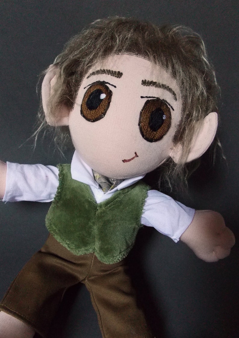 May include: A handmade doll with a fabric face, large brown eyes, and light brown hair. The doll wears a white shirt, a green vest, and brown pants. The doll has large, pointed ears.