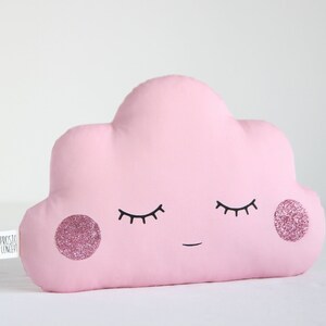 Puede incluir: Una almohada en forma de nube rosa con una cara sonriente, ojos cerrados y detalles de purpurina rosa. La almohada está hecha de tela suave y es perfecta para agregar un toque de fantasía a cualquier habitación.