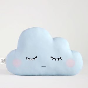 Puede incluir: Una almohada en forma de nube de color azul claro con una cara sonriente y los ojos cerrados. La almohada tiene mejillas rosadas y está hecha de tela suave.
