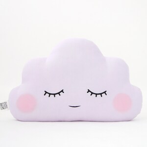 Puede incluir: Una almohada con forma de nube de color lavanda con una cara sonriente y los ojos cerrados. La almohada tiene mejillas rosadas y está hecha de tela suave.