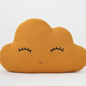 Puede incluir: Una almohada en forma de nube de color amarillo mostaza con una cara sonriente y pestañas. La almohada está hecha de tela suave y es perfecta para agregar un toque de fantasía a cualquier habitación.