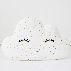 Puede incluir: Almohada blanca en forma de nube con lunares dorados y una cara sonriente con pestañas. La almohada tiene el texto "PROSTO CONCEPT" en la esquina inferior izquierda.