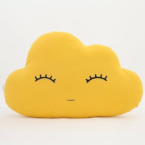 Puede incluir: Una almohada amarilla en forma de nube con una cara sonriente y pestañas bordadas.
