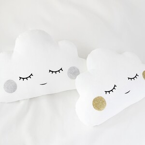 Puede incluir: Dos almohadas blancas en forma de nube con ojos y bocas bordados. Una almohada tiene un círculo de purpurina plateado y la otra tiene un círculo de purpurina dorada.
