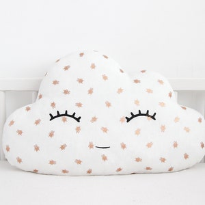 Puede incluir: Almohada blanca en forma de nube con una cara sonriente y bichos de purpurina dorada. La almohada tiene pestañas negras y una pequeña sonrisa.
