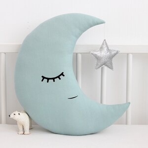 May include: A light blue crescent moon shaped pillow with a black embroidered eye and a silver glitter star hanging from the top.