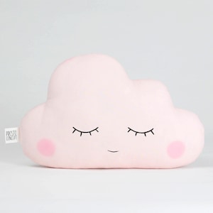 May include: A pink cloud-shaped pillow with a smiling face and closed eyes. The pillow has a small pink blush on each cheek.