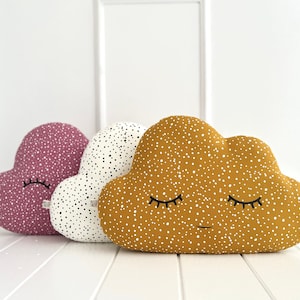 Puede incluir: Tres almohadas de felpa con forma de nube en rosa, blanco con puntos negros y amarillo con puntos blancos. Cada almohada tiene una cara sonriente con los ojos cerrados.