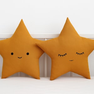 Puede incluir: Dos almohadas en forma de estrella de color amarillo mostaza con caras bordadas. Una almohada tiene una cara sonriente con ojos negros y una boca. La otra almohada tiene una cara dormida con los ojos cerrados y una pequeña sonrisa.