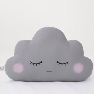 Puede incluir: Una almohada gris en forma de nube con una cara sonriente y los ojos cerrados. La almohada tiene mejillas rosadas.