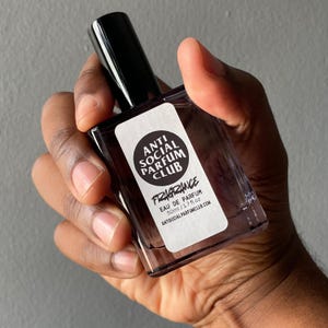 Peut inclure: Un flacon de parfum rectangulaire avec un bouchon noir est tenu dans une main. Le flacon porte une &eacute;tiquette blanche avec le texte "ANTI SOCIAL PARFUM CLUB" et "FRAGRANCE". Le flacon contient 50 ml de parfum.