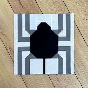 May include: A square fabric quilt block featuring a central black spider silhouette. The spider is constructed from pieced fabric segments in shades of white, light gray, and dark gray, creating a geometric, stylized arachnid design.