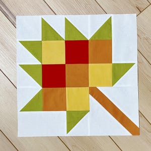 May include: A square fabric quilt block featuring a stylized leaf design constructed from geometric patches in shades of red, orange, yellow, and lime green, set against a white background with a diagonal orange stem extending from the corner.
