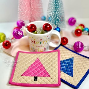 May include: A white ceramic mug with the text Merry and Bright in colorful letters, surrounded by a string of shiny red, pink, and blue ornaments. Two quilted fabric coasters are in the foreground, one with a pink tree and one with a blue tree.