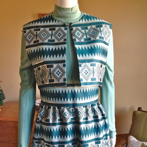 Op de afbeelding: Een teal en witte gepatroneerde jurk met een bijpassend teal en wit gepatroneerd vest. De jurk heeft lange mouwen en een hoge hals.
