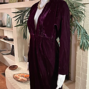 Puede incluir: Un vestido largo de terciopelo morado oscuro con mangas largas y escote pronunciado. El vestido está fruncido en la cintura y se exhibe en un maniquí. Se lleva un collar de cadena dorada con el vestido.