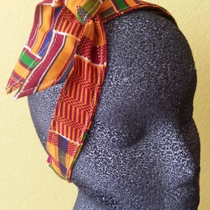 May include: A colorful headband with a bow, featuring a vibrant Kente cloth pattern. The fabric incorporates red, yellow, green, and blue geometric designs. The headband is displayed on a dark gray mannequin head against a yellow background.