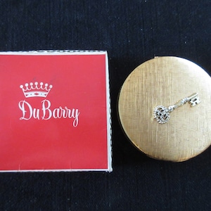 May include: A gold compact with a key design on the lid. The compact is in its original red box with the words "Du Barry" in white lettering.