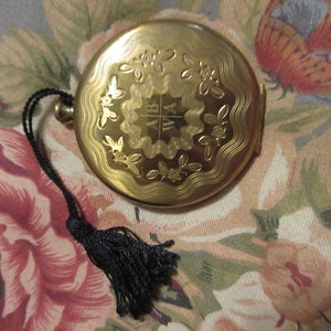 May include: A gold-colored compact with a black tassel. The compact has an engraved design with the letters "A B W A" in the center.