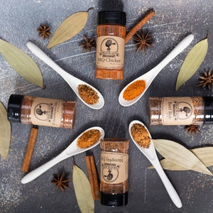May include: Five spice blends in glass jars with brown labels and black lids. The labels have a logo of a rooster and the text "Southern Flavors" and the name of the spice blend. The jars are arranged in a circle with white ceramic spoons filled with the spice blends. The spoons are arranged in a star shape. The background is a gray textured surface with cinnamon sticks, bay leaves, and star anise.