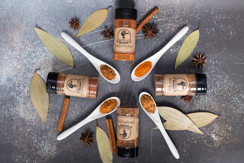 May include: Five spice blends in glass jars with brown labels and black lids. The labels have a logo of a rooster and the text "Southern Flavors" and the name of the spice blend. The jars are arranged in a circle with white ceramic spoons filled with the spice blends. The spoons are arranged in a star shape. The background is a gray textured surface with cinnamon sticks, bay leaves, and star anise.
