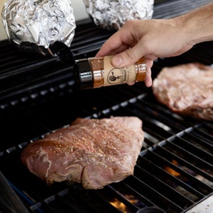 May include: A person is seasoning two pieces of meat on a black grill with a brown spice bottle labeled "The Little Butcher Shop".