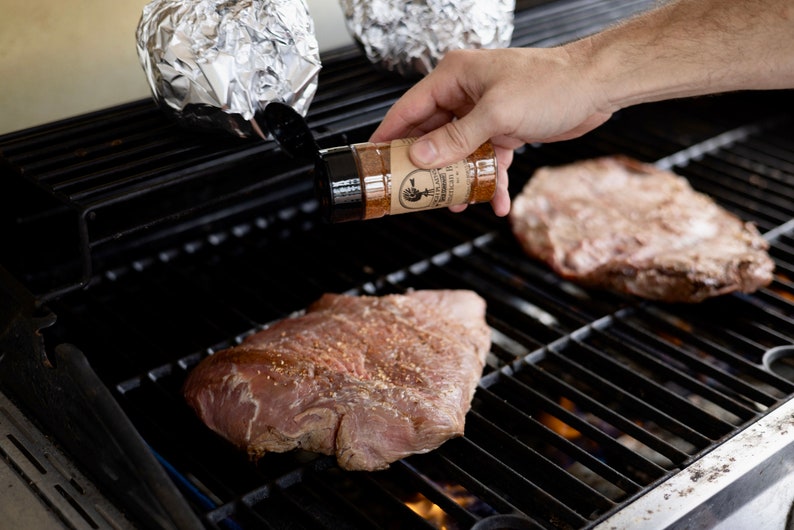 May include: A person is seasoning two pieces of meat on a black grill with a brown spice bottle labeled "The Little Butcher Shop".