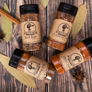 May include: Four glass jars with brown paper labels containing different spice rubs. The labels feature a windmill logo and the text "High Plains Spice Company". The jars are labeled "Beef Rub", "All American BBQ", "Memphis Rub", and "BBQ Chicken".