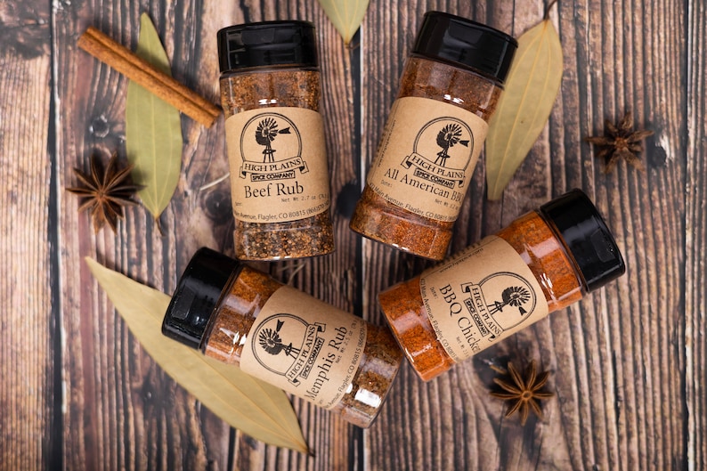 May include: Four glass jars with brown paper labels containing different spice rubs. The labels feature a windmill logo and the text "High Plains Spice Company". The jars are labeled "Beef Rub", "All American BBQ", "Memphis Rub", and "BBQ Chicken".