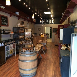 May include: A small shop interior with wooden shelves stocked with jars of spices, oils, and vinegars. The shop has a rustic, farmhouse aesthetic with exposed wooden beams and a barrel in the center of the room. The sign above the door reads "HPSC".