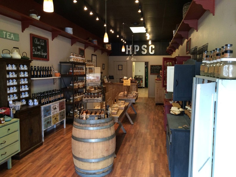 May include: A small shop interior with wooden shelves stocked with jars of spices, oils, and vinegars. The shop has a rustic, farmhouse aesthetic with exposed wooden beams and a barrel in the center of the room. The sign above the door reads "HPSC".
