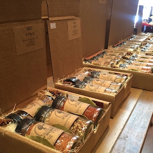 May include: A close-up of several cardboard boxes filled with small glass jars of spices. The boxes are labeled with different spice blends, including "BBQ 4-Pack" and "Hot Mix".