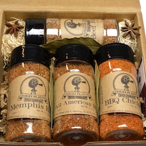 May include: A set of four spice jars in a brown cardboard box. The jars are filled with different colored spices and have brown labels with the text "High Plains Spice Company" and the names of the spices: "Memphis Rub", "All American BBQ", "BBQ Chick", and "Beef Rub".