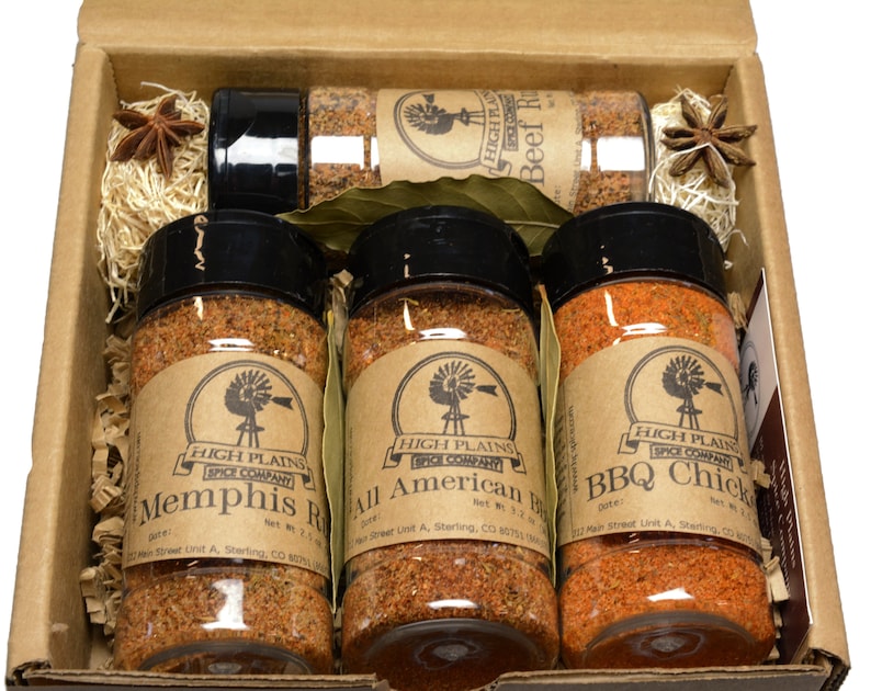 May include: A set of four spice jars in a brown cardboard box. The jars are filled with different colored spices and have brown labels with the text "High Plains Spice Company" and the names of the spices: "Memphis Rub", "All American BBQ", "BBQ Chick", and "Beef Rub".