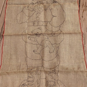 May include: A burlap canvas with a black line drawing of Santa Claus holding a teddy bear. Santa is wearing a hat, a belt, and boots.