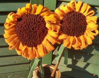 May include: Two felt sunflowers with brown centers in a beige ceramic vase. The vase is sitting on a white crocheted doily.
