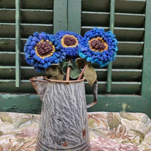 May include: A metal pitcher with a gray and white mottled finish and rust accents, containing three handcrafted felt flowers. The flowers feature layered blue petals, dark brown centers, and green felt leaves on thin stems.