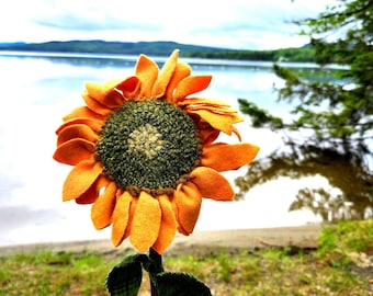 May include: A handcrafted sunflower with bright orange felt petals and a textured dark green center. The flower features a thin brown stem with two dark green leaves and is placed inside a clear glass bottle wrapped in thin black wire.