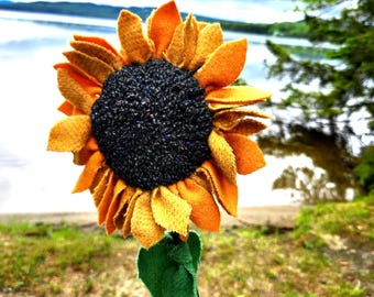 May include: A handcrafted sunflower decoration featuring layered orange and yellow felt petals surrounding a textured dark gray center. The flower is attached to a thin stem with a single green felt leaf and is placed in a clear glass bottle.